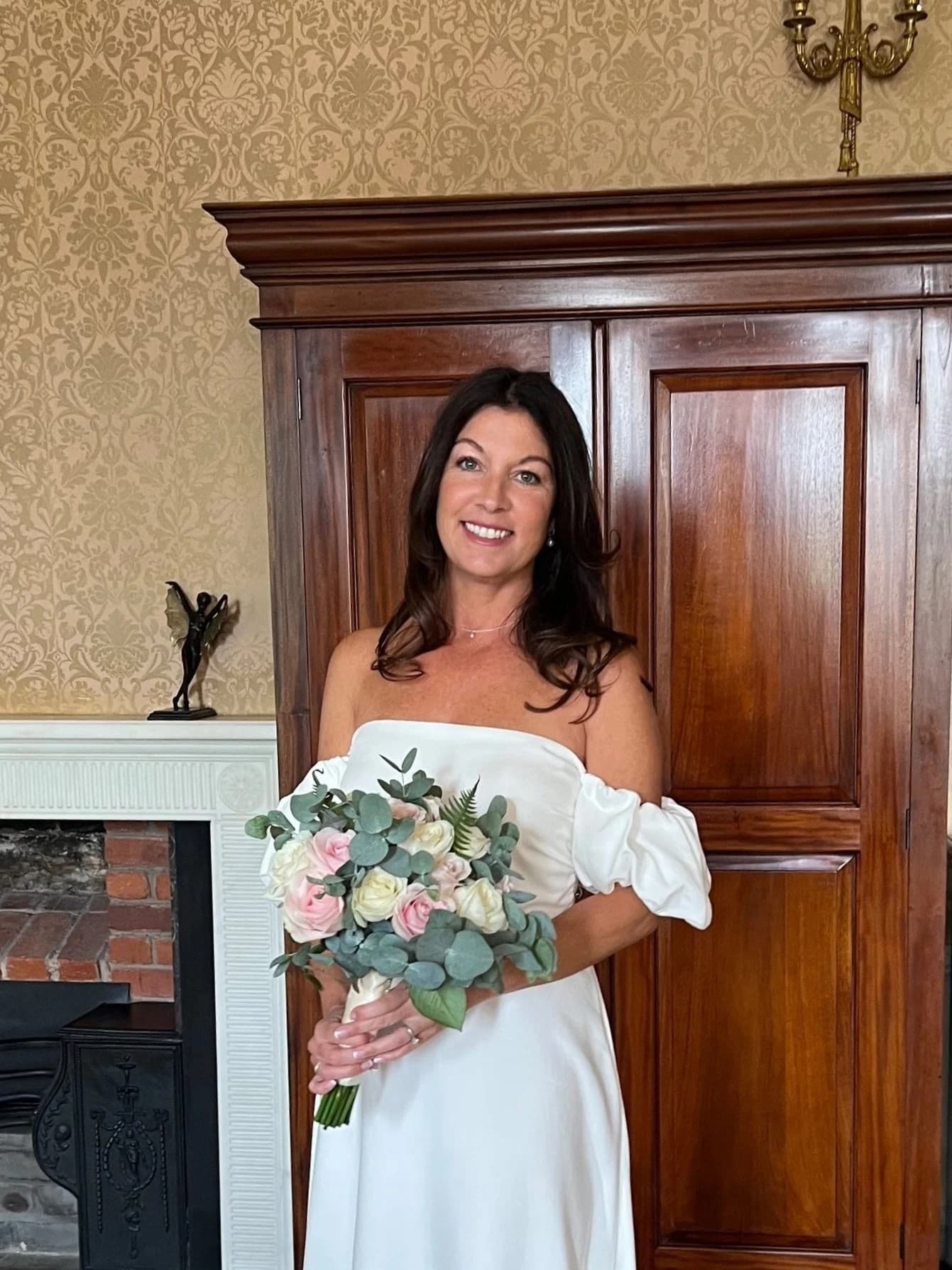 Bride holding a pink and white bouquet in front of a dark wooden wardrobe