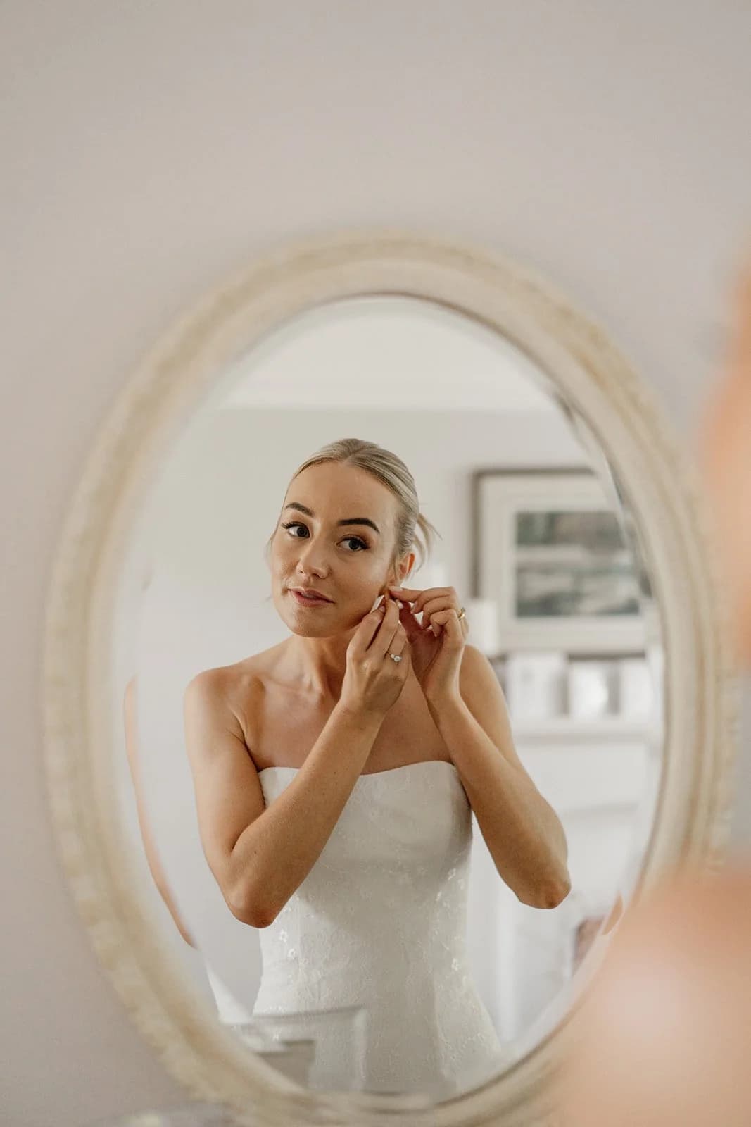 Bride fixing an earring, reflected in an oval mirror