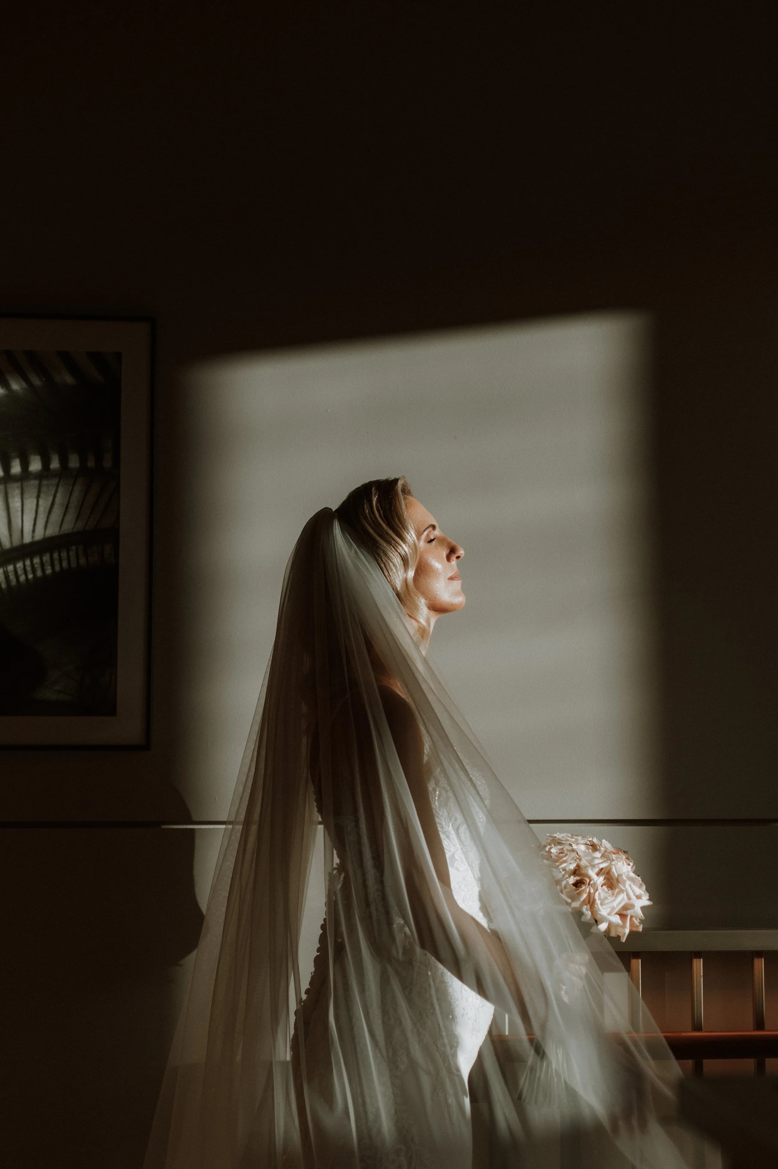 Bride in profile in a shaft of window light, eyes closed, long veil and a bouquet of blush roses