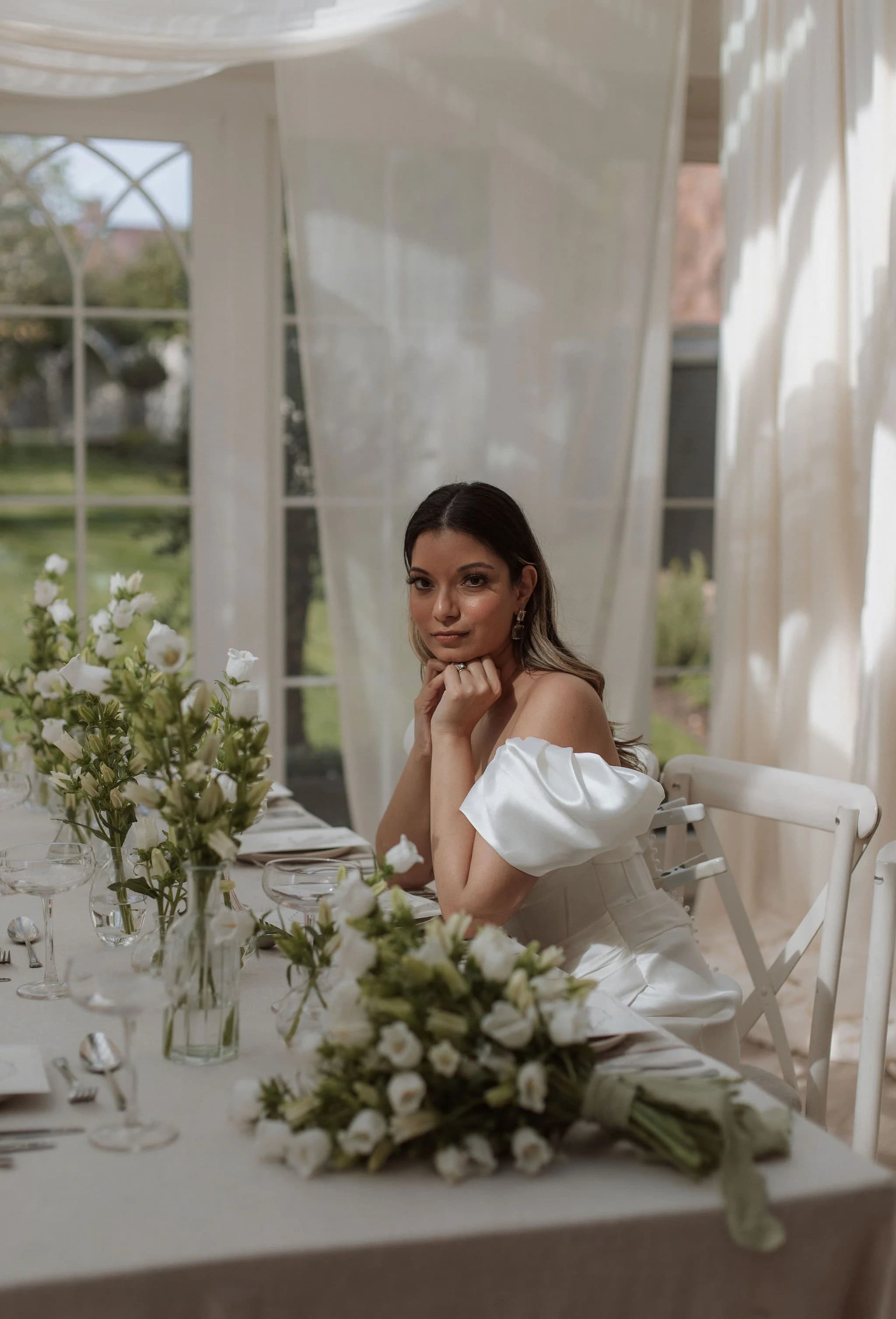 Bride in an off-the-shoulder satin dress at a table laid with white flowers, chin resting on her hand, lit from a window