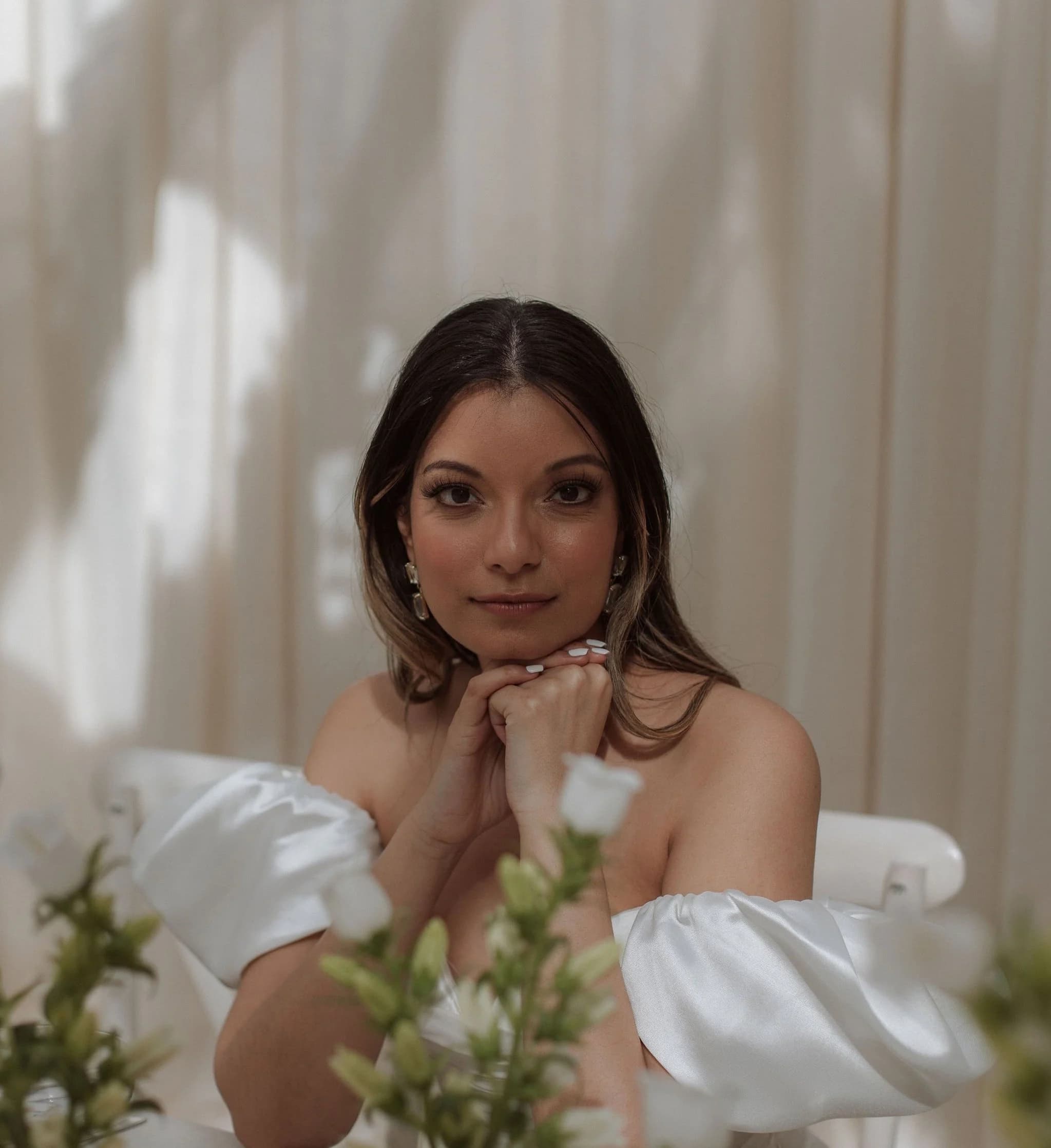 Bride with glowing, natural makeup resting her chin on her hands, white flowers in front of her