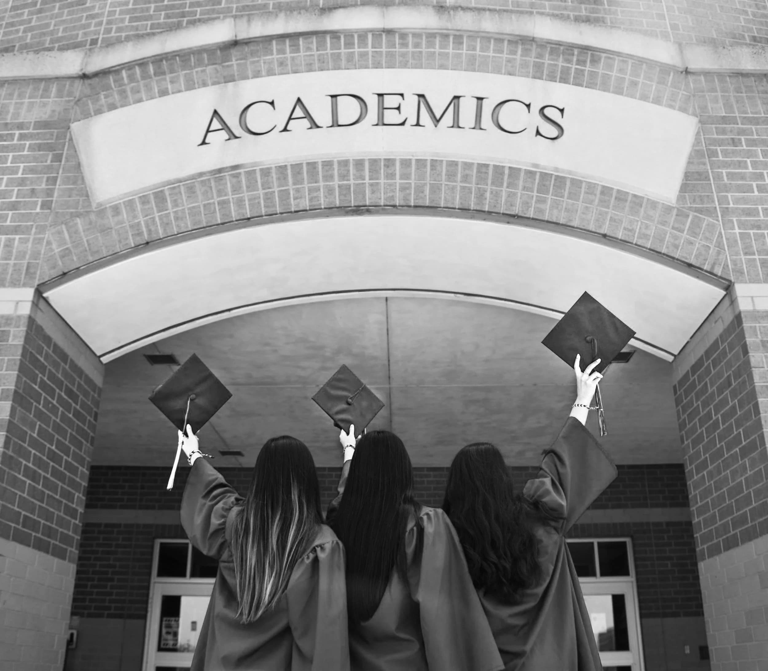 Three graduates throwing their caps under a sign reading Academics, in black and white