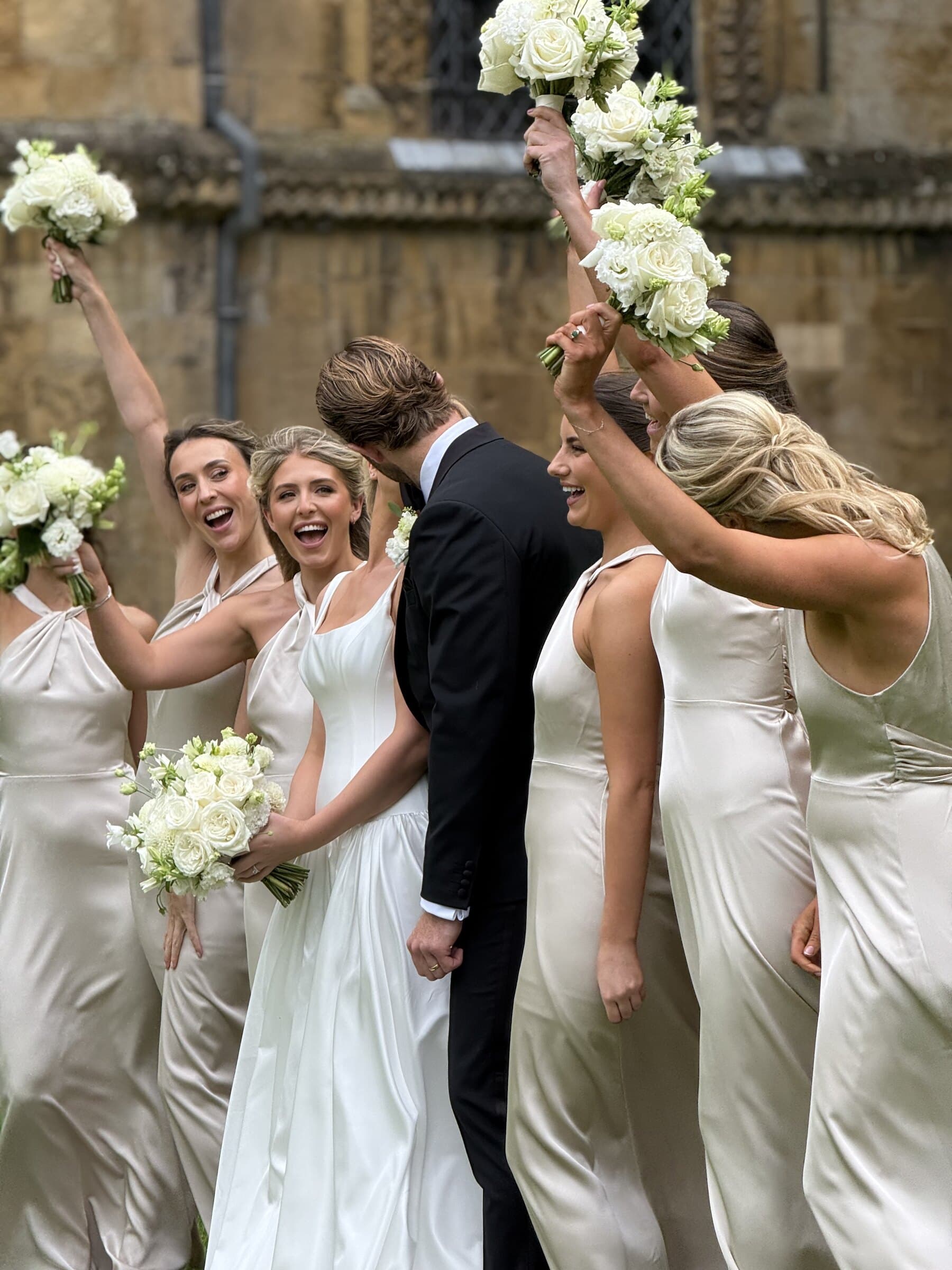 Bridesmaids in champagne satin raising their bouquets outside a stone church