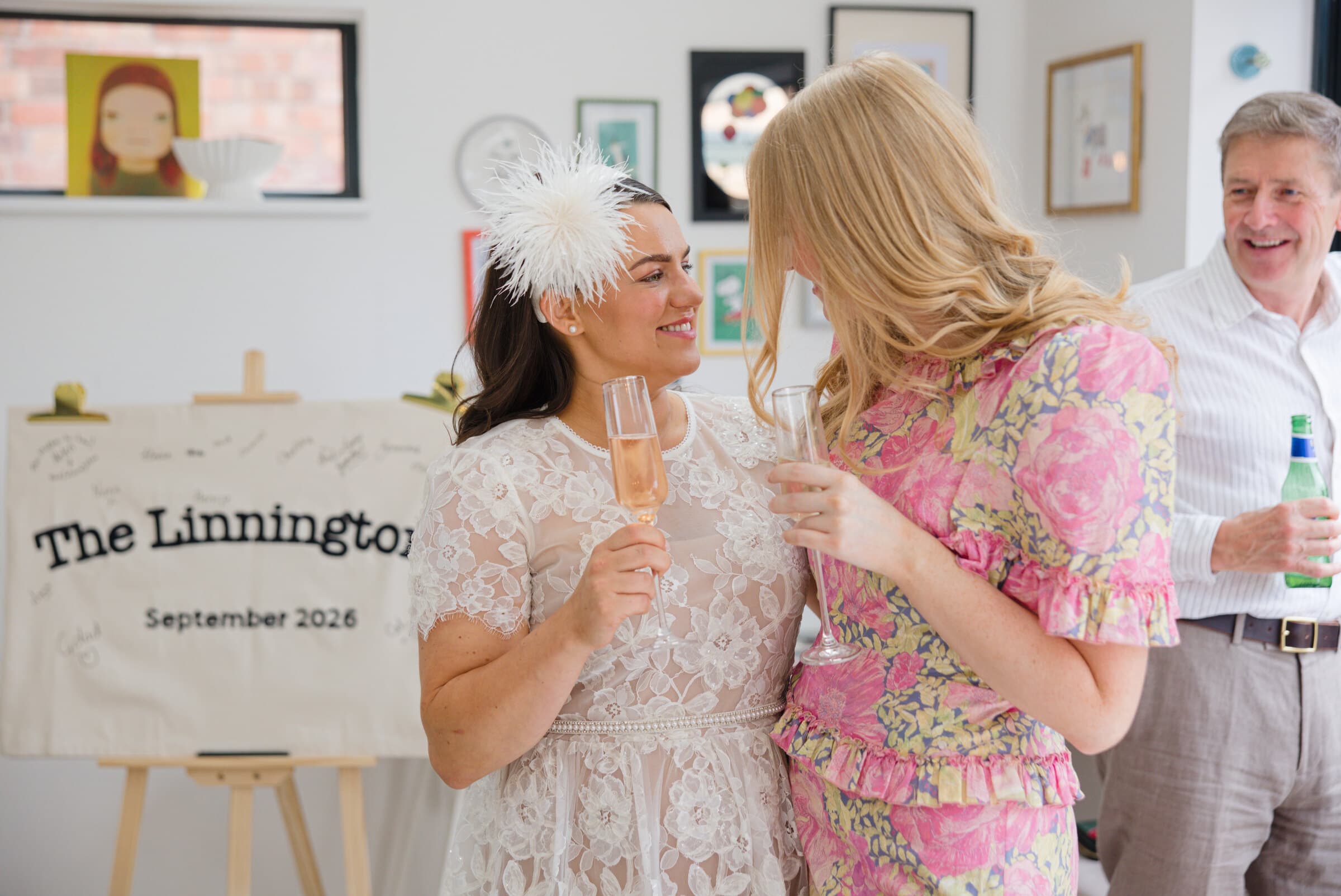 A bride in lace and a feather headpiece raising a glass with a friend in a floral dress