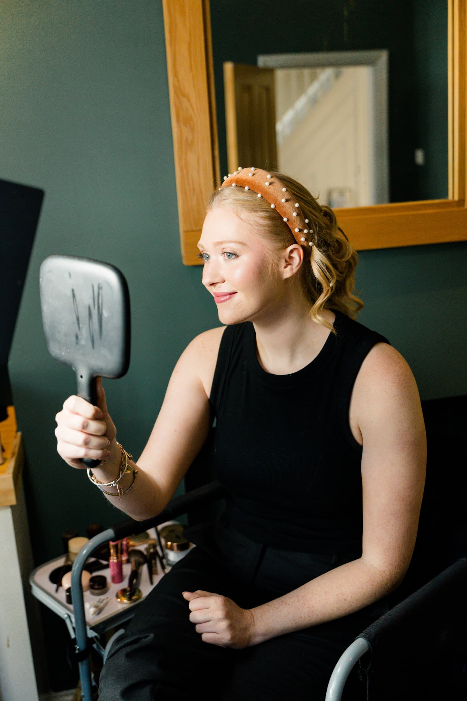 A guest in a pearl headband checking her makeup in a hand mirror beside the makeup kit