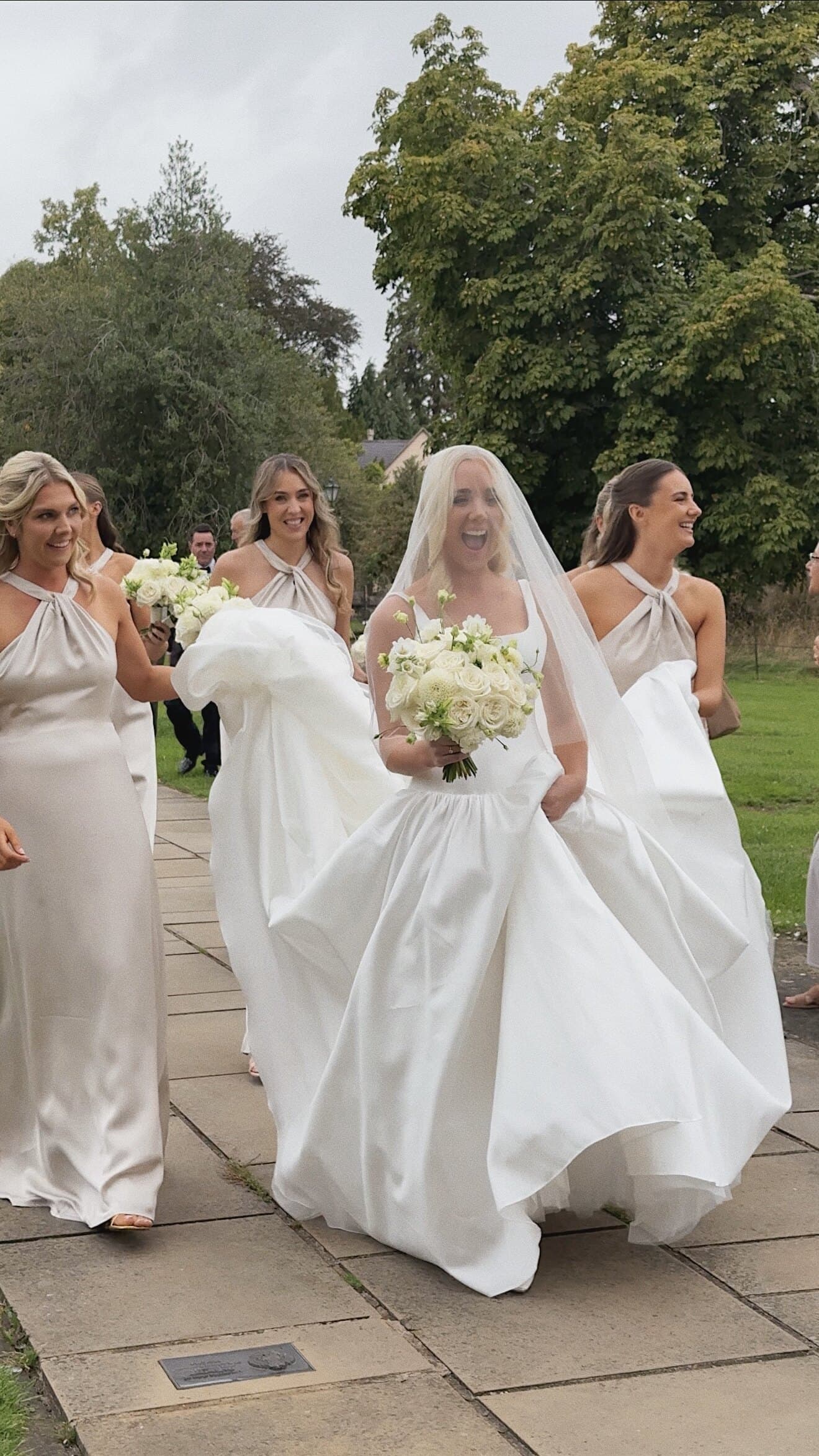 A bride laughing under her veil as her bridesmaids carry her train across the lawn
